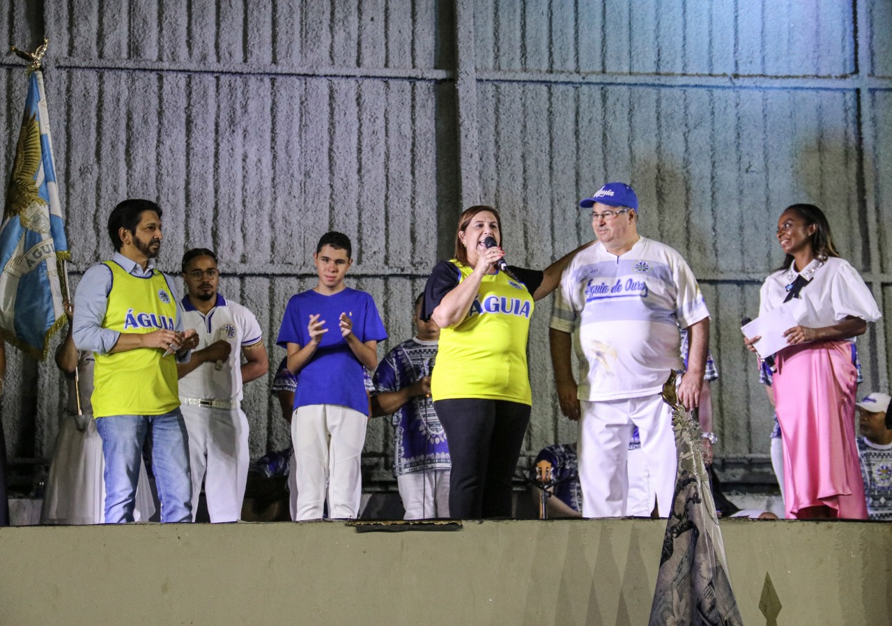 Na foto, que foi tirada do alto, é possível ter uma visão geral da quadra da Águia de Ouro. A quadra está cheia de integrantes da escola que participam do ensaio. No centro há passistas, mestre sala e porta bandeira, também é possível visualizar no canto esquerdo parte da bateria. Todos vestem roupas nas cores da escola: azul e branco. Ao fundo, há um palco com os intérpretes da escola, ao lado um grande telão que está exibindo o vídeo Samba com as Mãos. Na imagem, a intérprete de Libras traduz o samba-enredo da escola Águia de Ouro